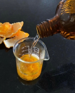 Becherglas mit geriebenen Mandarinenschalen und eine braune Flasche, die Alkohol zufügt.