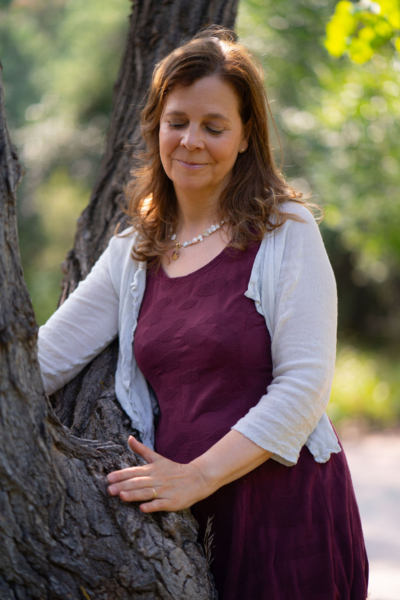 Fraue in einem weinroten Kleid mit weisser Jacke lehnt an einen Baumstamm und berührt die Rinde