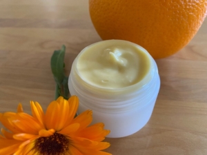 Salbendose mit cremiger Boybutter und eine Ringelblume im Vordergrund, hinten eine Orange
